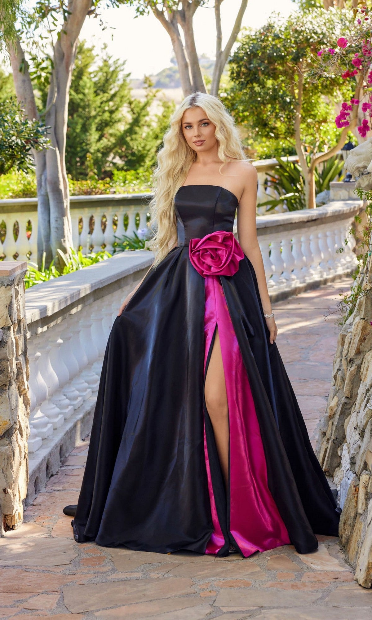 Wearing the Juliet JT2733H, a strapless black long prom dress with a pink rose accent and pink inner lining, a woman stands on a stone pathway surrounded by greenery.